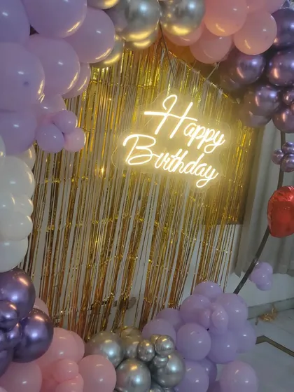 A beautiful birthday balloon arch in pastel purple, pink, and chrome silver, set against a gold foil curtain with a "Happy Birthday" neon sign.