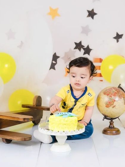 A baby boy sits with his cake in a travel-themed setup, complete with a vintage airplane and globe. A perfect theme for a little adventurer.