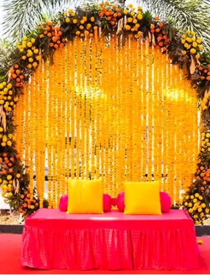 A vibrant outdoor Haldi photo booth with a circular backdrop of marigold strings. The bright pink seating provides a beautiful pop of color against the yellow flowers.