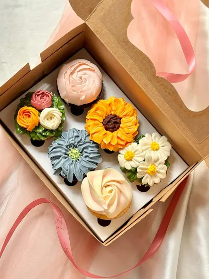 A gift box of six cupcakes decorated with intricate, multi-toned buttercream flowers. The collection displays a variety of floral piping techniques, including a sunflower, daisies, a rose, and other blossoms, perfect for elegant events like birthdays or anniversaries.