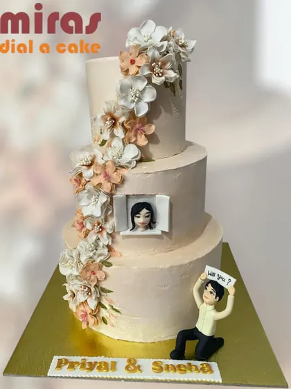 A cake that tells a love story. This elegant, three-tiered engagement cake captures a beautiful proposal moment, complete with a custom figurine, a photo of the happy couple, and a cascade of delicate, handmade sugar flowers.