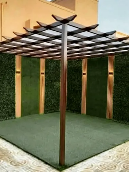 A square pergola with a distinctive single-post design and a grid roof. The back walls are covered in artificial turf and wood panels with integrated lighting, creating a modern, enclosed feel.