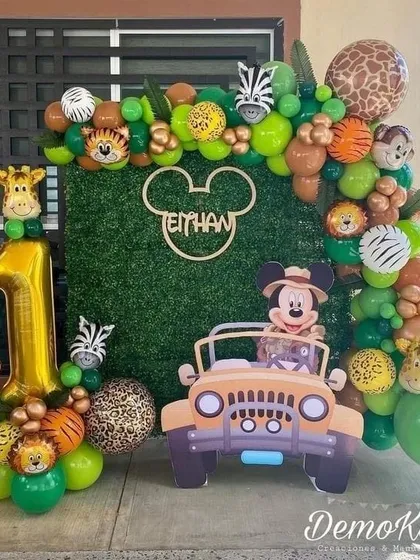 A unique Mickey Mouse safari theme. This outdoor setup combines a green hedge backdrop with a jungle balloon arch, a safari jeep prop with Mickey Mouse, and a large gold '1' for a first birthday.