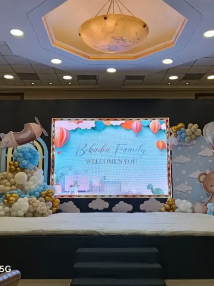 A grand stage setup featuring a hot air balloon and airplane theme. This design is perfect for a large baby shower or welcome party in a banquet hall, complete with a digital screen for custom messages and adorable teddy bear props.
