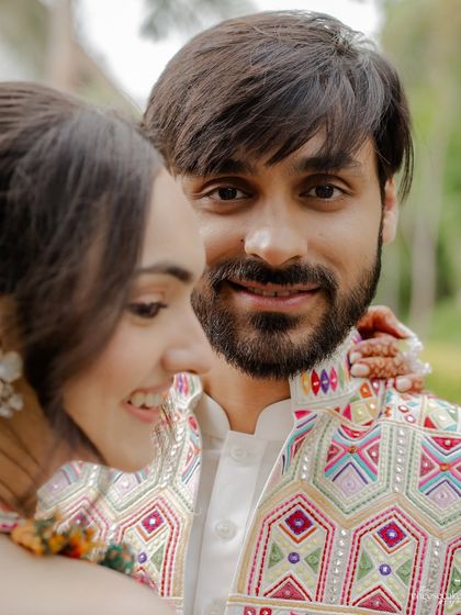 A playful and intimate shot where the groom is in focus, smiling at the camera, while his bride smiles beside him.