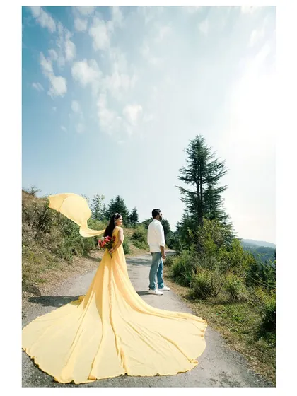 A woman in a vibrant yellow gown walks along a mountain road, its long train flowing behind her, creating a dramatic and colorful pre-wedding photograph.