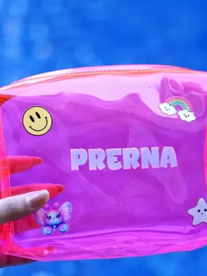 A close-up of a bright pink personalized jelly pouch. It's customized with the name 'Prerna' and decorated with cute patches like a smiley face, a rainbow, and a butterfly.