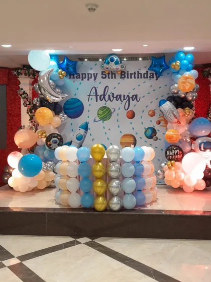 The main stage for a spectacular space-themed 5th birthday party. The setup features a personalized backdrop with planets and stars, surrounded by an elaborate balloon garland with rocket and astronaut accents.