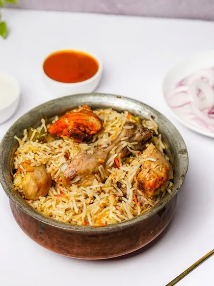 A traditional handi of chicken biryani, garnished and served with sides of raita, mint chutney, and pickled onions. The clean, white background and classic props make this a perfect shot for delivery app menus like Zomato or Swiggy.