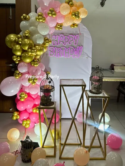 An elegant indoor birthday setup featuring a pink, white, and gold balloon garland on an arched backdrop. We added a 'Happy Birthday' neon sign and decorative props like birdcages on pedestals to enhance the theme.