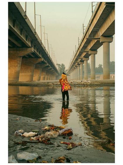 A person carries a religious idol into the polluted waters of the Yamuna, with two metro bridges looming in the background. The image is a stark commentary on faith, pollution, and urban development.