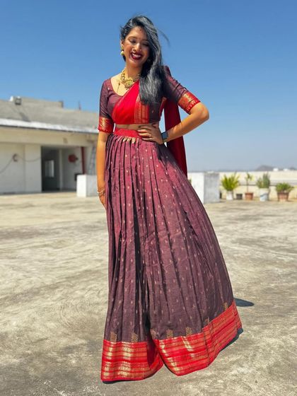 This half-saree features a deep maroon skirt with a traditional gold and red border, paired with a contrasting red blouse and dupatta for a vibrant, celebratory look.
