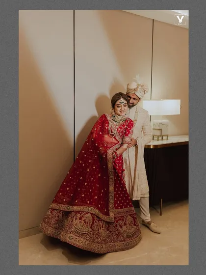 A full-length portrait of the couple in a hotel room, the bride's red lehenga adding a vibrant pop of color. The soft lighting and their relaxed pose create an intimate and elegant feel.