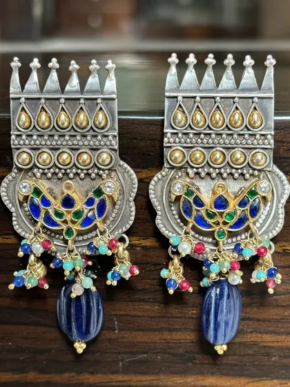These statement fusion earrings are designed to turn heads. They feature a crown-like silver top, gold-plated accents, colourful enamel peacock motifs, and are finished with dangling blue beads.