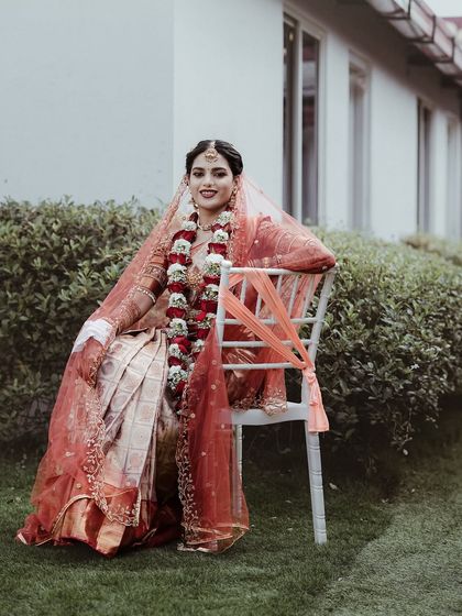 A full-length portrait of the bride, seated elegantly outdoors. Her beautiful red and gold saree, garland, and serene expression are the focus of this classic shot.