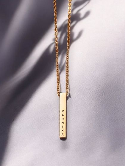 A clean, minimalist shot of a gold bar pendant engraved with 'Varnika'. The soft lighting and simple background focus all attention on the personalized piece.