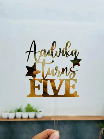 The "Aadvika Turns FIVE" topper held up against a bright background, showing off the clean cuts and reflective quality of the gold acrylic.