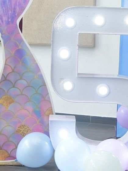 A close-up of the details from a mermaid-themed party. This shot features a large, light-up marquee number '5' next to a custom-printed mermaid tail prop, surrounded by pastel balloons.