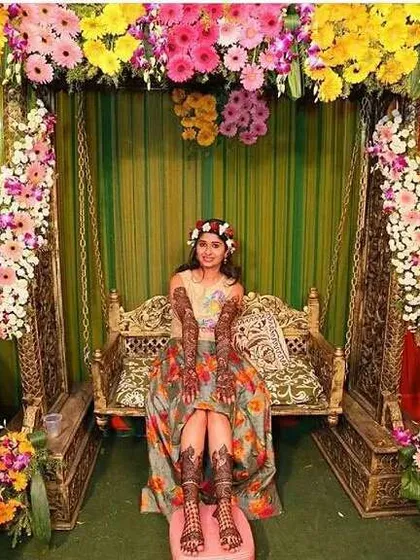 A beautiful Mehandi setup with a bride on a decorated jhula. The background is filled with colorful flowers like gerberas, creating a perfect photo moment.
