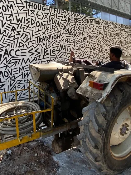 A man on a tractor in front of the art-covered boundary wall of MAP, a picture of the museum meeting the street.