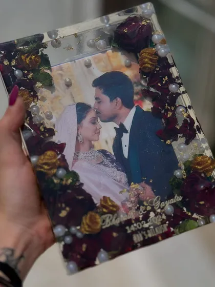 A close-up of a rectangular photo frame, preserving deep red and yellow roses with pearls. The couple's photo captures a tender moment, now frozen in time.