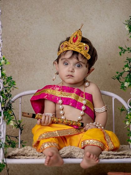 A toddler dressed as Krishna, sitting on a decorated swing. His wide-eyed, slightly surprised expression is a perfect example of a precious, candid moment.