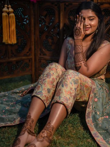 A happy bride showing off her simple and elegant mehendi for a pre-wedding event. The design is focused on the palms and feet, leaving the arms open.