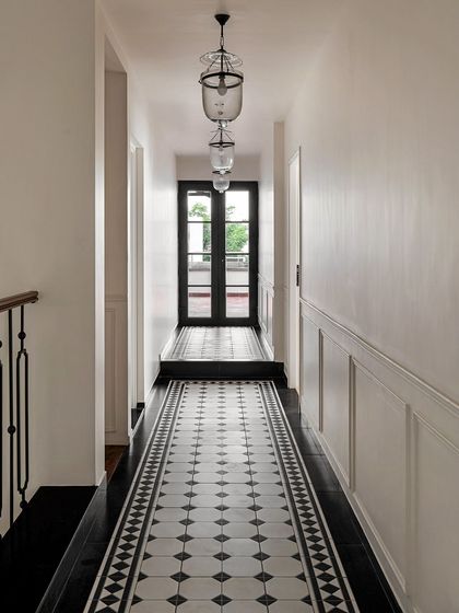 A long corridor featuring classic black and white patterned floor tiles and simple wall paneling. The repetition of pendant lights draws the eye forward, creating a sense of length and timeless elegance.