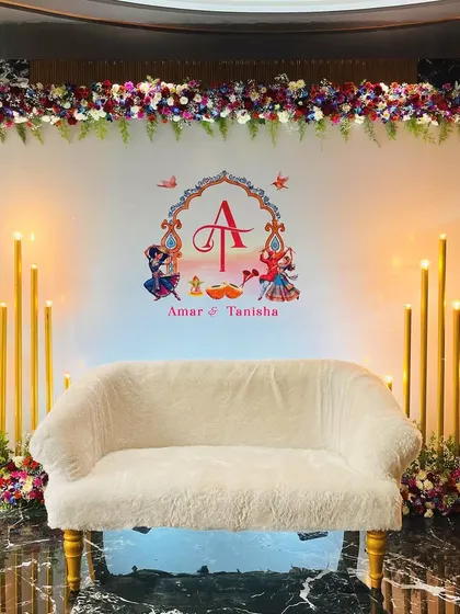 A simple and elegant stage for a Godhana and Chunadi ceremony. The white backdrop features a custom couple's monogram with traditional motifs, framed by floral arrangements and tall candles.