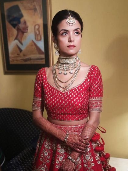 A full-view of a bride in her stunning red lehenga, showcasing her complete look. The makeup is balanced to complement the heavy, multi-layered necklace and intricate outfit.