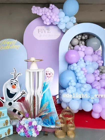A magical Frozen-themed birthday party setup perfect for any fan of Elsa and Olaf. This decoration features custom arched backdrops in pastel blues and purples, a cascading balloon garland, and life-sized character cutouts to complete the winter wonderland scene.