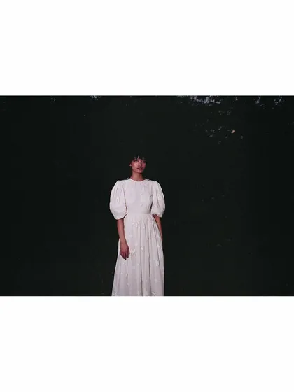 An atmospheric shot for the Sruti Dalmia campaign. The model is styled in a white dress with puff sleeves, positioned against a dark, natural background to create a moody and cinematic image that emphasizes solitude and form.