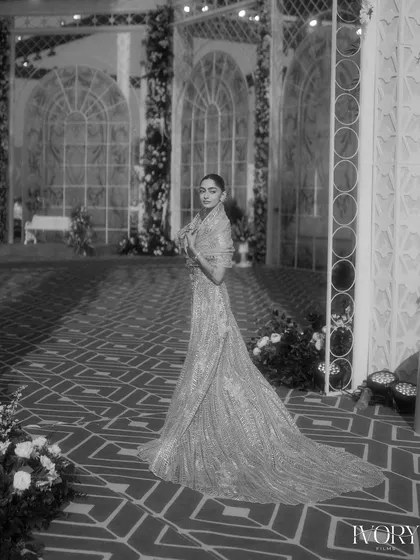 A full-length black and white portrait of the bride at her reception. The grand, arched windows and geometric floor create a stunning, high-fashion backdrop.