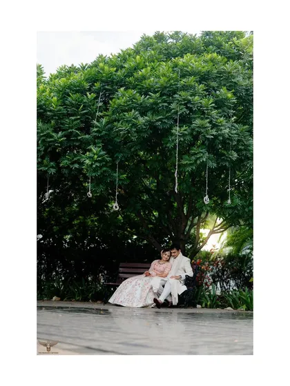 A quiet, romantic scene of a couple sitting together on a bench under a large, leafy tree. This wide shot captures the peacefulness of the moment and the feeling of finding your own world together.