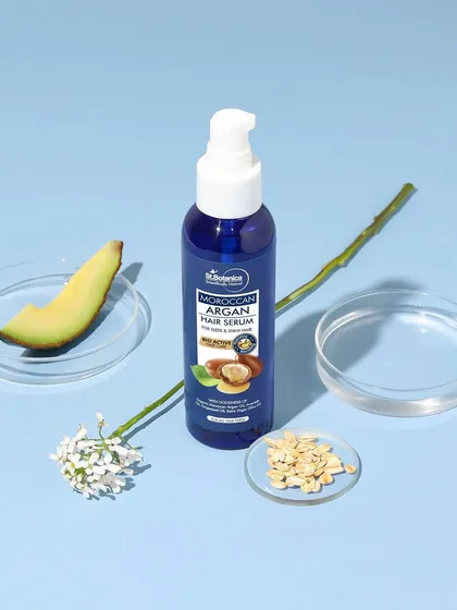 A clean and minimal composition for a hair serum. The product is styled with an avocado, a flower, and lab glassware to highlight its blend of natural ingredients and scientific formulation.