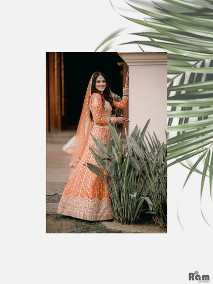 A full-length shot of the bride in her vibrant orange lehenga, framed by lush green leaves. This adds a touch of nature and artistry to her engagement day portraits.