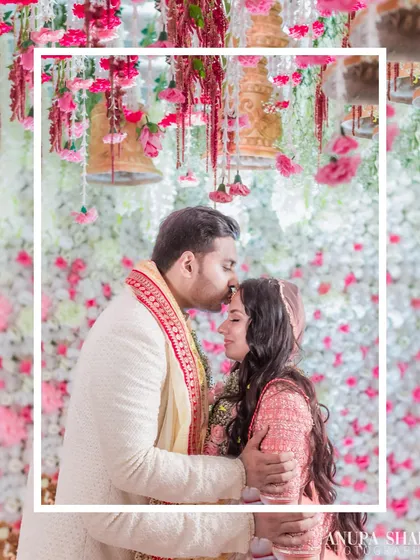 A beautiful portrait of the groom kissing his bride's forehead, framed by a stunning floral decor of hanging bells and flowers. The composition creates a sense of being in a romantic wonderland.