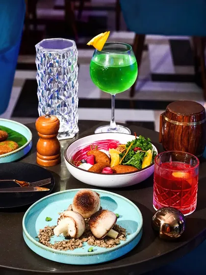 A table spread in a lounge, featuring appetizers like stuffed mushrooms and a bright green cocktail. The image captures the trendy and energetic vibe of the location.