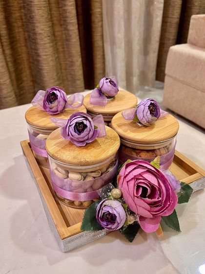 A modern take on gifting dry fruits. This set includes five glass jars with wooden lids, filled with different nuts and decorated with purple ribbons and flowers, all presented on a sleek silver tray.