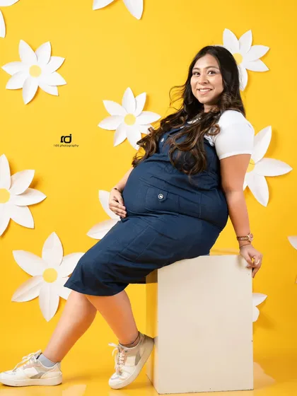 A bright and happy solo portrait. The cheerful yellow background and daisy props perfectly match the joyful smile of the mom-to-be in her casual denim outfit.