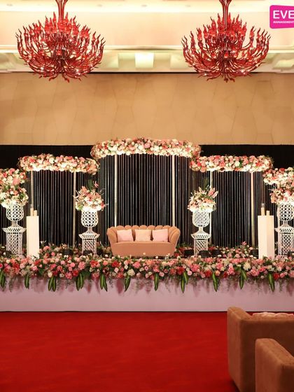 A sophisticated reception stage with a modern black-paneled backdrop. The design is elevated by arrangements of pink and white florals in classic white urns, creating a chic and timeless look.
