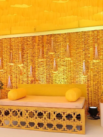 For this indoor haldi ceremony, I created a complete yellow theme. The backdrop is made of woven genda phool, with hanging floral tassels, and the seating is a simple wooden bench, creating a clean and modern look.