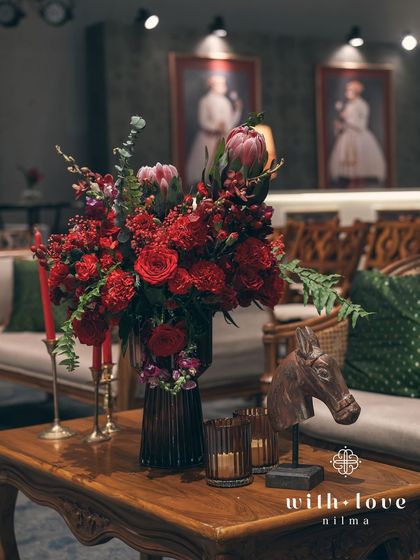 A rich floral arrangement of red roses and proteas sits on a classic wooden table. In the background, framed Raja Ravi Varma paintings and deep green walls create an atmosphere of timeless elegance and artistic flair.