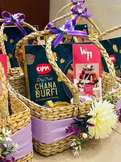 A beautifully curated return gift hamper for a wedding or engagement. This wicker basket features a box of Om Sweets' Dry Fruit Chana Burfi, adorned with a lavender ribbon and delicate flowers, offering a tasteful thank you to guests.