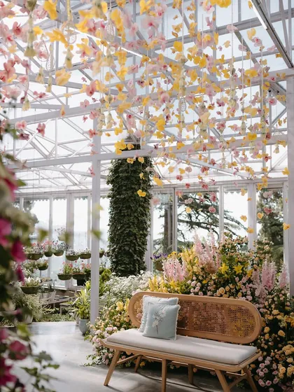 Another view of the stunning seating area inside the greenhouse. The rattan bench is surrounded by a lush garden of flowers, with the delicate floral canopy creating a magical, filtered light from above.