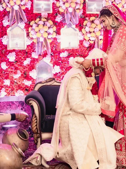 A classic Varmala moment captured from a low angle. The groom bows to receive the garland from his bride against a backdrop of red and white flowers, a traditional pose that I capture with an eye for composition and emotion.