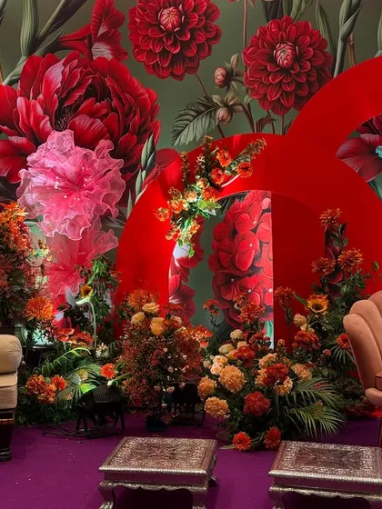 A colorful and eclectic seating arrangement for a pre-wedding event. The backdrop features a bold red floral wallpaper and large red arches, surrounded by vibrant floral arrangements and unique fabric flowers.