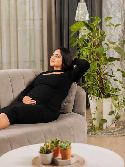 A moment of peaceful daydreaming. The mother-to-be relaxes on the couch in her living room, surrounded by plants, creating a calm and natural lifestyle portrait.