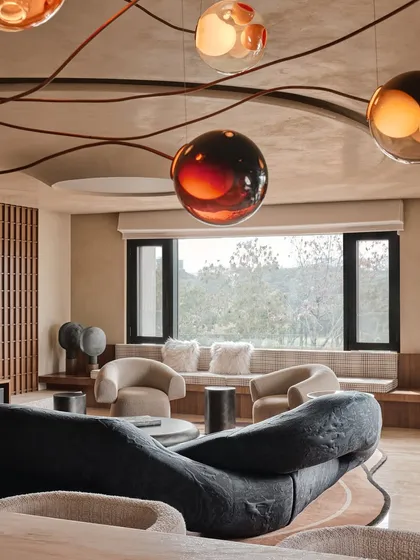 The living room, with its unique, branching chandelier that spreads across the ceiling. The large windows connect the space to the outdoors, while the organic shapes of the furniture create a comfortable, modern lounge.
