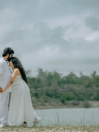 A serene and romantic portrait of a couple in matching white outfits, holding hands by a calm lake. This style of pre-wedding photography is timeless and elegant.